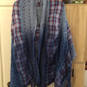 May include: A blue and red plaid shawl with a lace trim. The shawl is hanging on a wooden hanger. The lace trim is a darker shade of blue than the plaid fabric. The shawl is draped over a wooden cabinet.