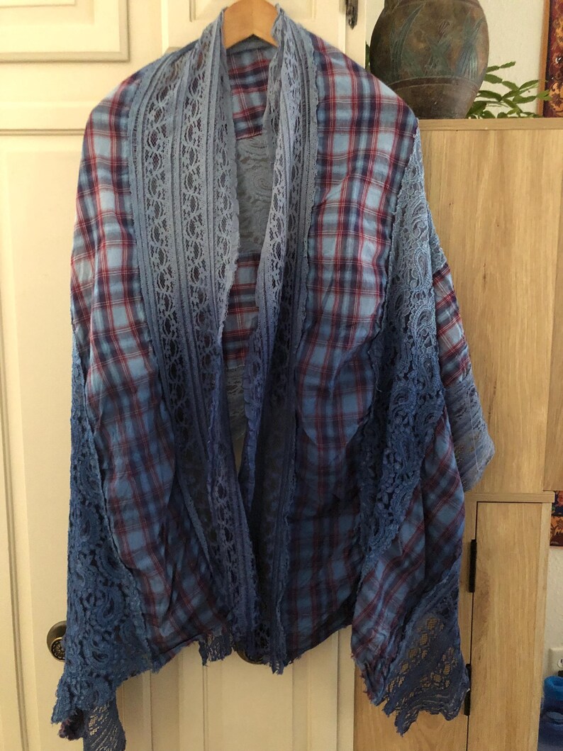 May include: A blue and red plaid shawl with a lace trim. The shawl is hanging on a wooden hanger. The lace trim is a darker shade of blue than the plaid fabric. The shawl is draped over a wooden cabinet.