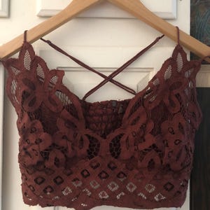 May include: A burgundy lace bralette with a floral design and crisscross straps. The bralette is hanging on a wooden hanger against a white background. The lace has a detailed pattern.