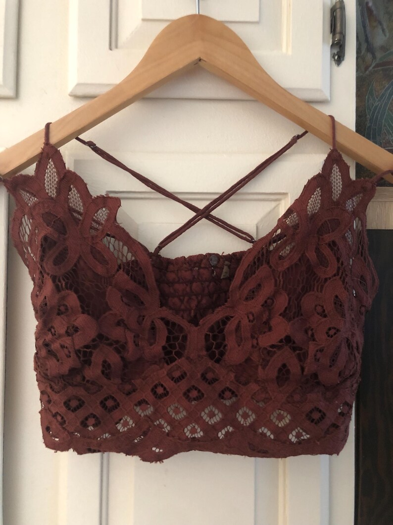 May include: A burgundy lace bralette with a floral design and crisscross straps. The bralette is hanging on a wooden hanger against a white background. The lace has a detailed pattern.