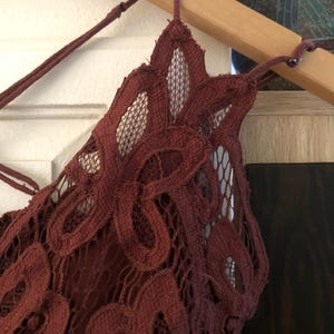 May include: A burgundy-colored lace top with a delicate floral pattern. The top features a halter-style neckline and is hanging on a wooden hanger. The intricate lace design is visible, showcasing the texture and detail of the garment.