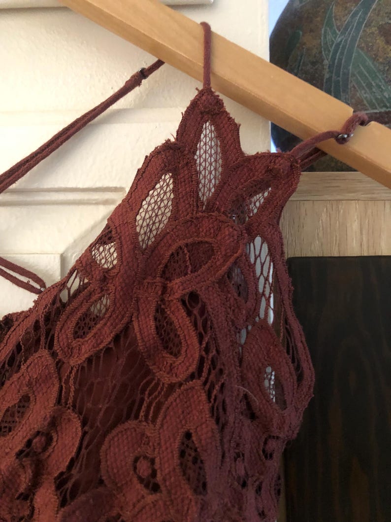 May include: A burgundy-colored lace top with a delicate floral pattern. The top features a halter-style neckline and is hanging on a wooden hanger. The intricate lace design is visible, showcasing the texture and detail of the garment.