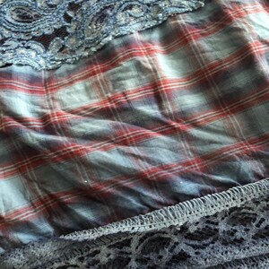 May include: Close-up of fabric layers featuring a blue lace overlay, a red and blue plaid pattern, and a dark blue lace trim. The textures and colors create a layered effect, suitable for apparel or craft projects.