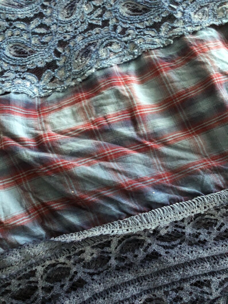 May include: Close-up of fabric layers featuring a blue lace overlay, a red and blue plaid pattern, and a dark blue lace trim. The textures and colors create a layered effect, suitable for apparel or craft projects.