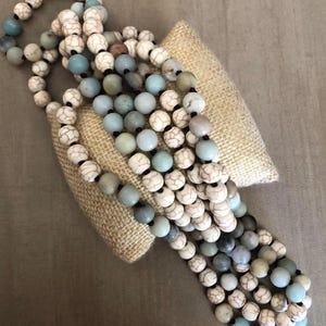 May include: A long beaded necklace featuring a repeating pattern of round white beads with dark brown veining and smooth, matte-finish light blue and grey beads. The beads are strung on a dark cord, creating a multi-layered look with varied textures.