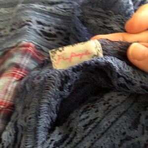 May include: Close-up of a blue, open-weave fabric with a tag that reads "Free People" in pink script. The fabric appears to be layered over a red and white plaid pattern. The texture of the fabric is visible.