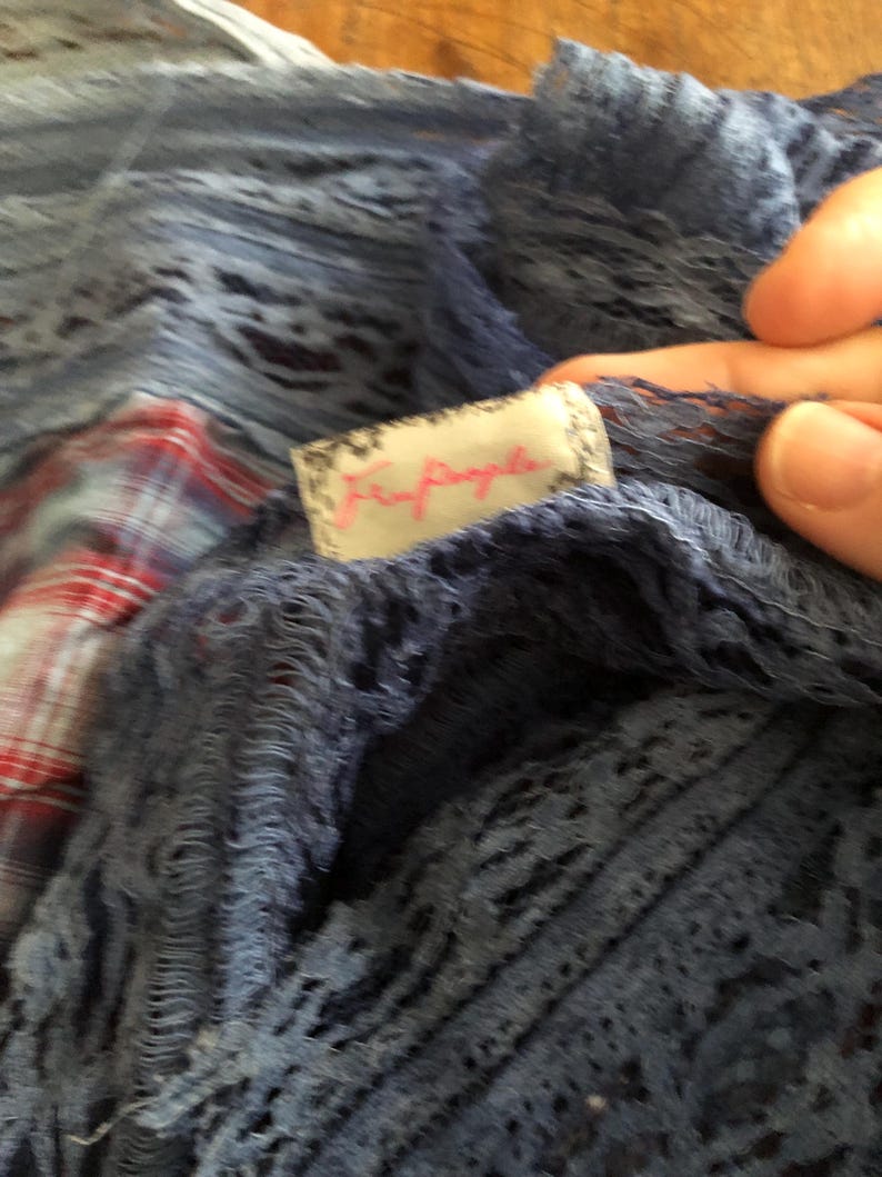 May include: Close-up of a blue, open-weave fabric with a tag that reads "Free People" in pink script. The fabric appears to be layered over a red and white plaid pattern. The texture of the fabric is visible.
