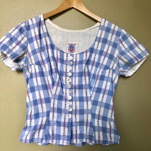 May include: A vintage blue and white plaid blouse with short sleeves and a scoop neckline. The blouse features a row of white buttons down the front and vertical floral accents. The garment is hanging on a wooden hanger.