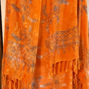 May include: A bright orange scarf featuring a tie-dye effect and a repeating pattern of grey sea turtles. The fabric includes a horizontal band with geometric diamond and triangular motifs across the lower section, finished with long fringe.