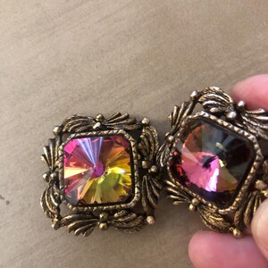 Puede incluir: Un par de pendientes cuadrados vintage con cristales iridiscentes multicolores. Los cristales están engastados en un marco de metal color bronce con un diseño de hojas decorativas. Los pendientes están en la mano de una persona.