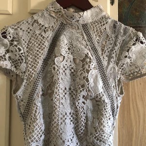 May include: A pale grey lace top with short sleeves and a high neckline. The intricate floral and geometric patterns create a delicate, openwork design. The top is hanging on a wooden hanger.