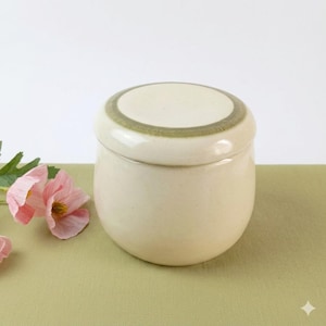 May include: A round, off-white ceramic jar with a matching lid. The lid features a thin, circular band of muted olive green around its edge. The jar has a smooth, rounded body and a glossy finish, presenting a simple and functional design.