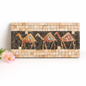May include: A rectangular mosaic art piece featuring three camels in warm tones of orange, red, and yellow. The camels are set against a dark background, framed by a border of beige and brown square tiles. The artwork is likely for home decor.