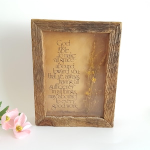 May include: A framed artwork with a rustic wooden frame. The artwork features a printed quote on a tan background, with dried flowers. The quote reads "God is Able to make all grace abound toward you..."