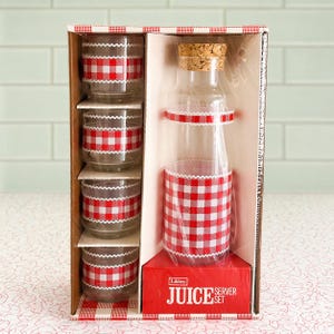 May include: A vintage juice server set in a cardboard box. The set includes a clear glass bottle with a cork stopper and four matching glasses. Each glass and the bottle are decorated with red and white gingham patterns. The box reads "JUICE SERVER SET".