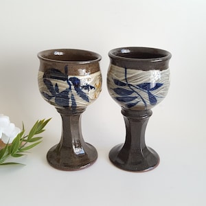 May include: Two handmade ceramic goblets. The goblets have a dark brown stem and rim, with a lighter brown and white body. The body of each goblet is decorated with blue leaf-like patterns. The goblets are displayed on a white surface.