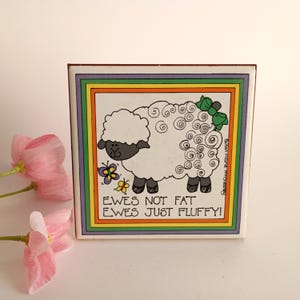 May include: Square ceramic tile featuring a cartoon sheep with a fluffy white body, black face and legs, and a green bow. The tile has a colorful border and text that reads "EWES NOT FAT EWES JUST FLUFFY!"