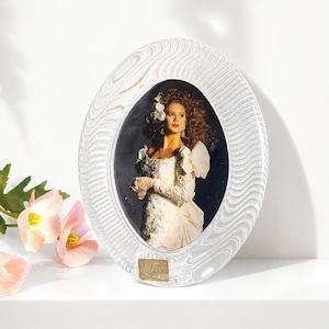 May include: Oval-shaped clear glass picture frame with a wavy, textured design. The frame holds a photograph of a woman in a white dress. The frame has a gold-colored label at the bottom that reads "MIKASA".