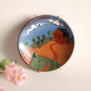 May include: A decorative ceramic plate with a hand-painted landscape scene. The plate features a red barn, trees, a winding path, and a blue sky with white clouds. The plate is mounted with gold-colored metal hangers.