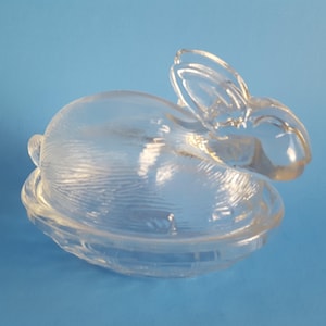 May include: A clear glass bunny-shaped dish with a lid. The bunny has long ears and a textured surface. The dish is sitting on a blue surface. This decorative dish is perfect for Easter.