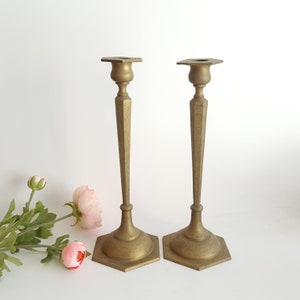 May include: Two antique brass candlesticks. Each candlestick has a hexagonal base, a long, tapered stem, and a square top. The brass has a warm, golden hue, and the stems are decorated with etched floral patterns. The candlesticks are displayed on a white surface.