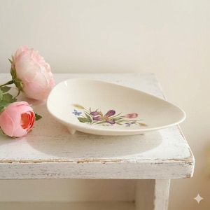 May include: Cream-colored, teardrop-shaped ceramic dish with a floral design featuring purple irises, blue accents, and green leaves. The dish rests on three small feet. Pink roses are in the background, set on a distressed white wooden surface.