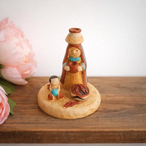 May include: A handcrafted ceramic figurine depicting a woman carrying a water jug on her head, with a child and a small bowl. The woman wears a blue shawl and a turquoise necklace. The figurine is on a tan base, with a wooden background.