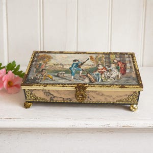 May include: A rectangular vintage jewelry box with a hinged lid. The lid features a detailed painting of people in a garden setting. The box has a marble-like pattern on the sides, gold trim, and decorative feet.