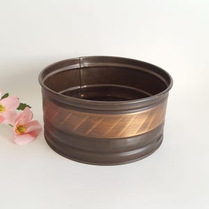 May include: A round, dark brown metal planter with a copper-colored band featuring a diagonal pattern. The planter has a simple, cylindrical shape and is likely used for displaying plants or flowers. The overall aesthetic is vintage or rustic.