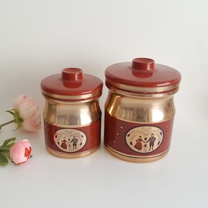 May include: Two matching vintage jars with lids. The jars have a reddish-brown exterior with gold-colored bands and bases. Each jar features a gold oval with a couple illustration. The jars are likely for kitchen storage.