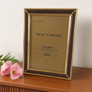 May include: A rectangular photo frame with a gold-colored border and a dark brown textured trim. The frame has the text "Tiffany Collection" and "Solid Brass" with dimensions of 5" x 7". The frame is on a wooden surface with pink flowers.