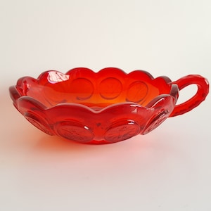 May include: A vibrant red glass bowl with a scalloped rim and a handle. The bowl features embossed circular designs around the edge. The glass is a deep, transparent red, and the bowl is likely vintage or antique.