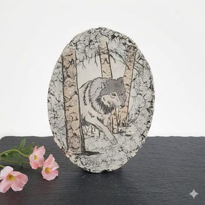 May include: Oval-shaped ceramic art piece featuring a wolf in a forest scene. The artwork depicts a gray wolf walking through birch trees. The piece has a textured, cracked surface with a neutral color palette.