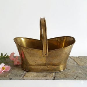 May include: A polished brass basket with an oval base and a single vertical handle attached to the center of the sides. The metal surface features a warm golden tone with subtle variations in texture and a smooth, reflective finish throughout.