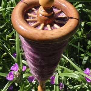 May include: A wooden drop spindle with a dark brown wheel-like top and a light brown base. The spindle is filled with a cone of purple yarn. The background features green grass and small purple flowers.
