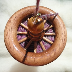 May include: A wooden drop spindle with a wheel-like design. The spindle features a polished, brown wooden wheel with spokes and a central wooden bead. The center is filled with purple and gray yarn, and a strand of yarn extends from the top.