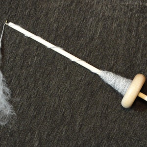 May include: A wooden drop spindle with a light grey yarn wrap and a wisp of white fibre attached. The spindle has a natural wood finish and is set against a dark grey background. This tool is used for hand spinning yarn.