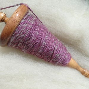 May include: A wooden drop spindle with a purple and gray yarn wrapped around the shaft. The spindle has a rounded top and a pointed bottom. The yarn is a mix of purple and gray fibers.