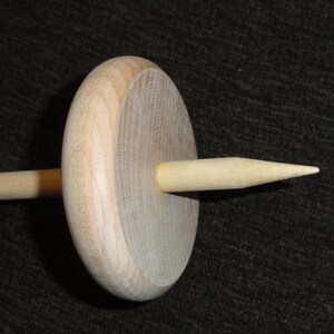 May include: A wooden drop spindle, a tool for hand spinning yarn. The spindle features a circular wooden whorl and a tapered shaft. The natural wood color is visible throughout the tool, with a smooth finish.