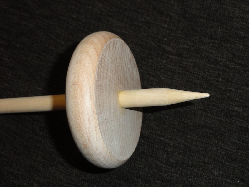 May include: A wooden drop spindle, a tool for hand spinning yarn. The spindle features a circular wooden whorl and a tapered shaft. The natural wood color is visible throughout the tool, with a smooth finish.