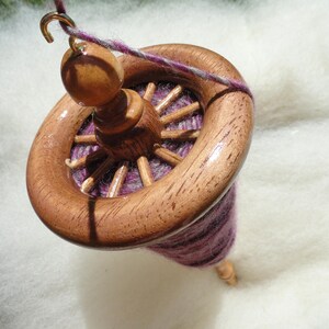May include: A wooden spinning wheel with a dark brown finish. The wheel has a circular design with spokes and a central spindle. The yarn is a mix of purple and gray. The wheel is suspended by a gold-colored hook.