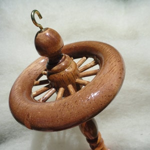 May include: A wooden drop spindle with a wheel-like whorl and a brass hook. The spindle is made of polished wood, showcasing the natural grain. The whorl has spokes, and the overall design is for spinning yarn.
