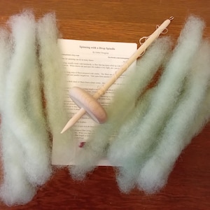 May include: A wooden drop spindle with a light-colored wooden whorl and a long, thin shaft, resting on a sheet of paper and surrounded by pale green wool roving. The paper has text that reads "Spinning with a Drop Spindle".