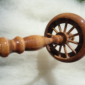 May include: A wooden spinning wheel hand spindle with a wheel-like design. The spindle is made of polished wood, featuring a wheel with spokes and a handle. The background is a soft, white material, suggesting a textile craft theme.