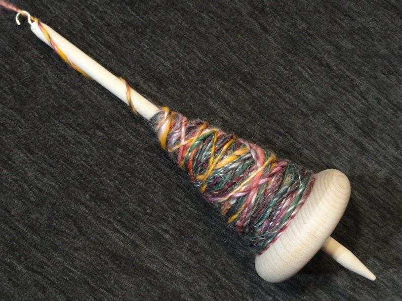 May include: A wooden drop spindle with a colorful yarn wrap. The spindle has a natural wood finish with a small hook at the top. The yarn is a mix of colors including pink, yellow, green, and brown.