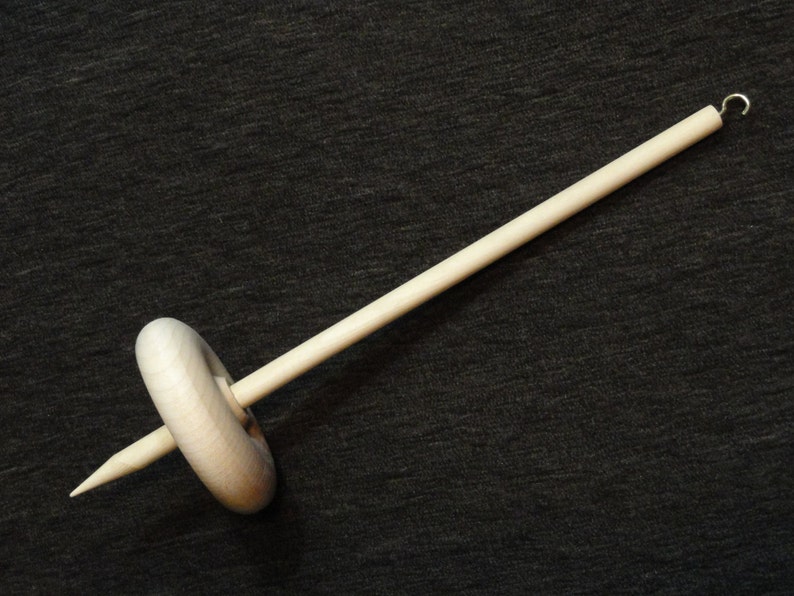 May include: A wooden drop spindle, a tool for hand spinning yarn. The spindle has a long, slender shaft with a pointed end and a small hook at the top. A wooden whorl is positioned in the middle of the shaft.