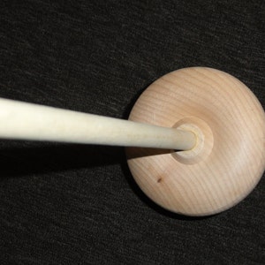 May include: A wooden spindle with a round base and a long, cylindrical shaft. The base is light-colored with visible wood grain. The shaft extends from the center of the base. The spindle is designed for spinning yarn or thread.