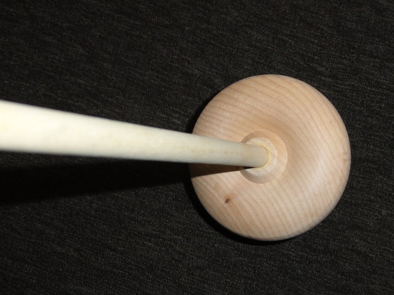 May include: A wooden spindle with a round base and a long, cylindrical shaft. The base is light-colored with visible wood grain. The shaft extends from the center of the base. The spindle is designed for spinning yarn or thread.