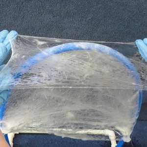 May include: A clear, thin, and flexible material is stretched over a blue, arched frame. The material appears to be held by gloved hands. The frame is supported by a white structure, and the background is a dark blue.