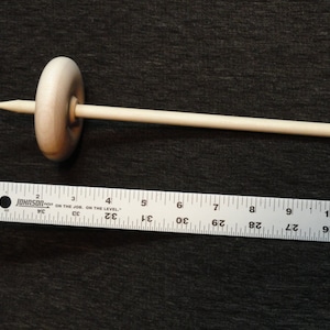 May include: A wooden drop spindle, a tool for hand spinning yarn. The spindle has a rounded whorl and a long, slender shaft. The shaft is approximately 10 inches long. The whorl is about 1.5 inches in diameter. The spindle is made of light-colored wood.